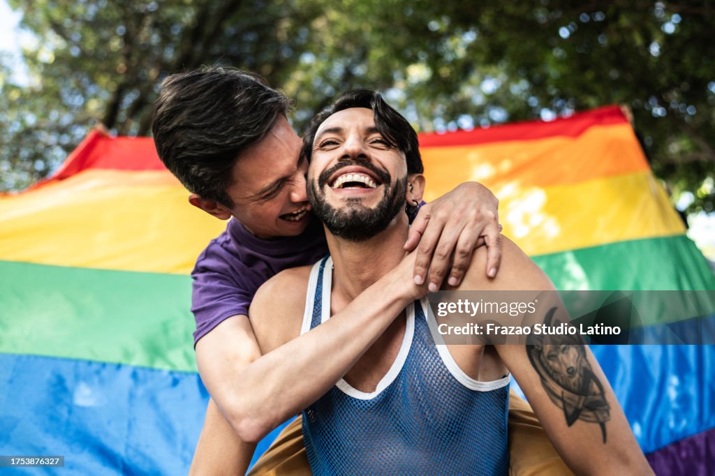 Couple gay piggyback à l’extérieur avec drapeau arc-en-ciel sur fond
