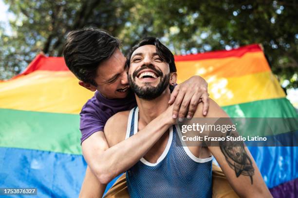 gay couple piggyback outdoors with rainbow flag on background - gay pride parade in colombia stock pictures, royalty-free photos & images