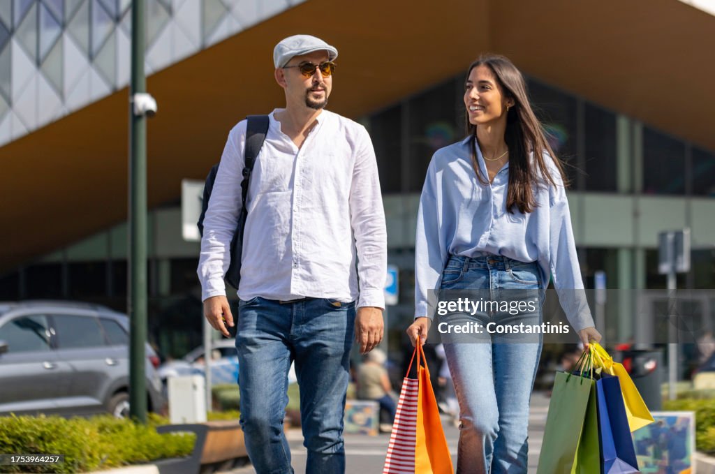Boyfriend and girlfriend, walking in front of the shopping mall, while a young woman carrying shopping bags