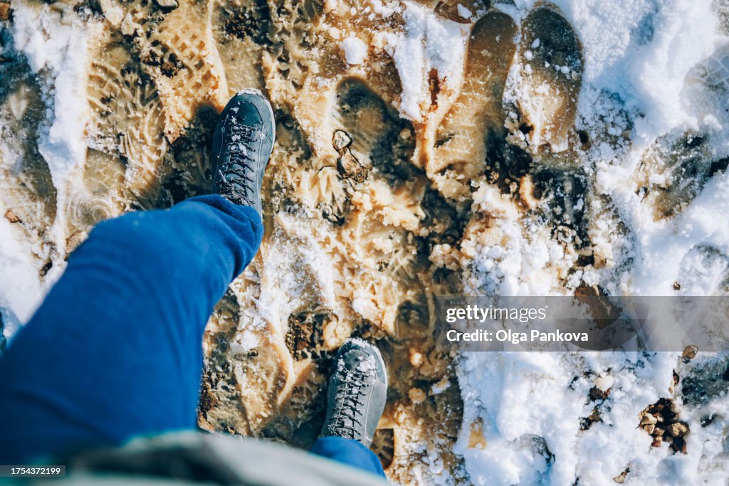 POV first-person view of feet with boots on the snow and a dirty road