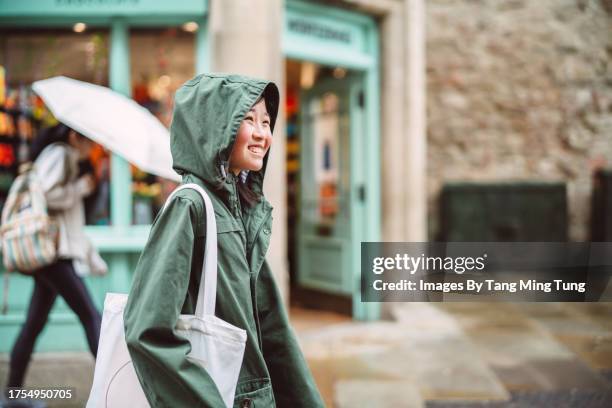 cheerful teenage girl in raincoat walking under the rain while exploring in a town - raincoat stock pictures, royalty-free photos & images