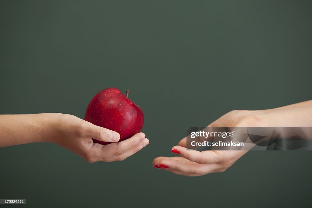 Little student hand giving red apple to teacher before blackboard