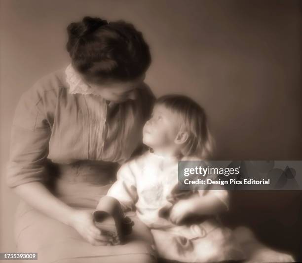 Mother sits with her toddler daughter and a holds a toy as they look at each other.