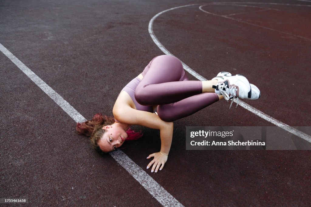 Sporty girl in fitness suit trains outside on playground. Fit young woman does handstand. Attractive brunette with long highlighted hair does exercises for balance on her hands on basketball court.