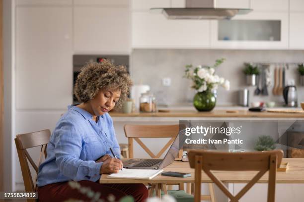 businesswoman taking notes while working on laptop at home office. - laptop kitchen table stock pictures, royalty-free photos & images