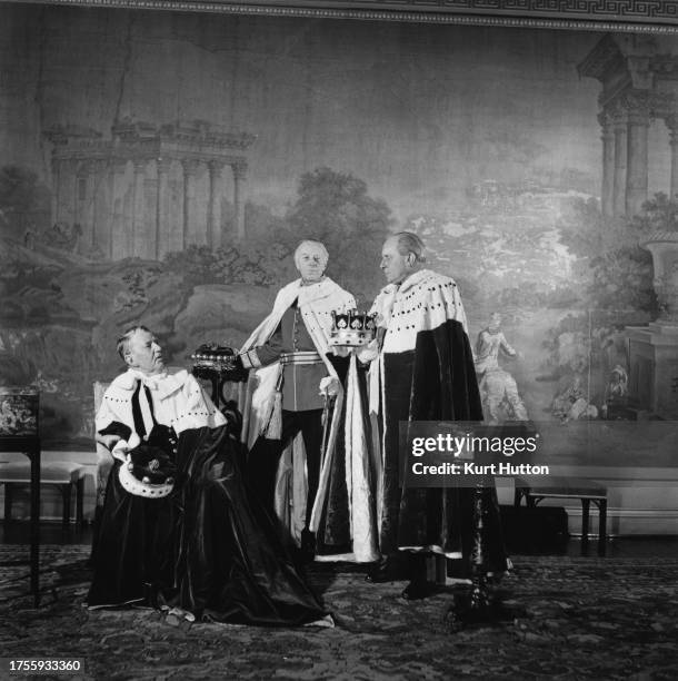 Three dignitaries each wearing ermine-trimmed ceremonial robes, posing with crowns before a large painting, during preparations for the Coronation of...