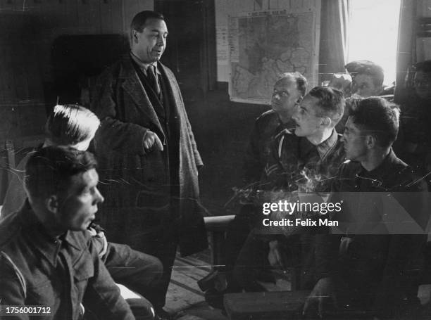 British writer and broadcaster JB Priestley stands speaking to British military personnel, a 'Daily Express War Map' on the wall in the background,...