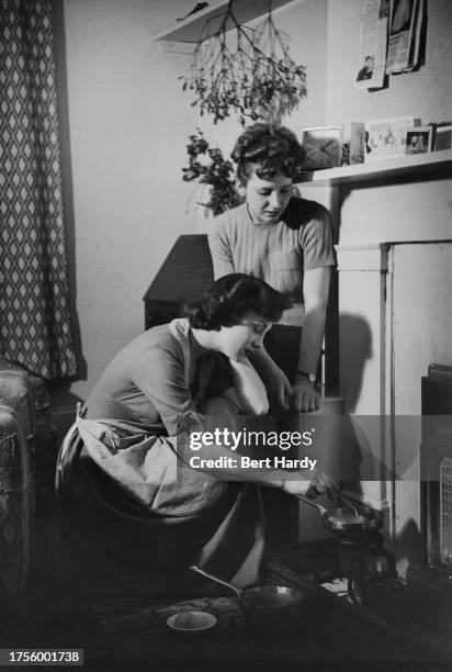 Two women with one stirring a pan as she sits beside a camping stove in the hearth of a fireplace, a small pan on the stove with a second pan on the...