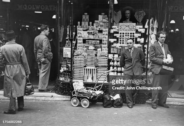 Four men, one standing beside two bags and a pushchair, outside a seafront souvenir shop, postcards, rock and sunglasses displayed in the window,...