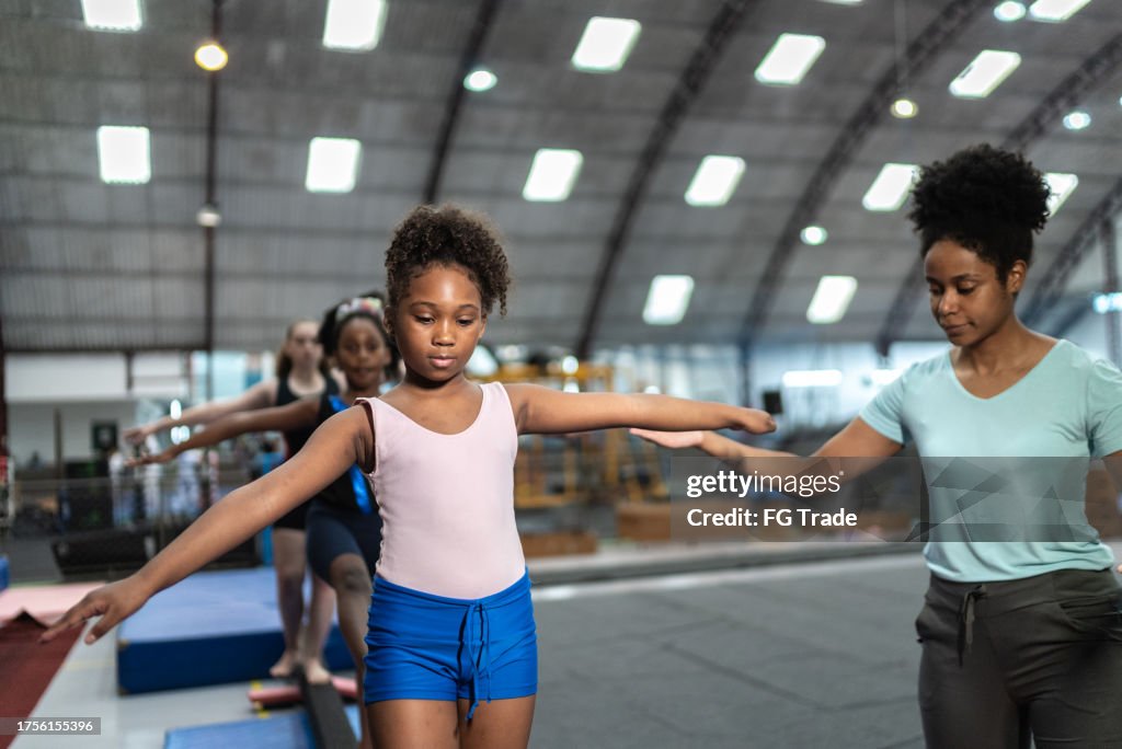 Girl being helped by her coach to walk on a balance beam in a gymnasium