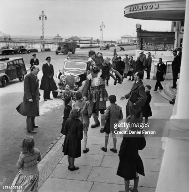 Man in costume holds a small child aloft, surrounded by children and men outside the entrance to a seafront ballroom, United Kingdom, 1952. Original...