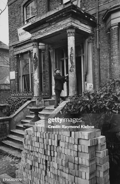 Man standing before the portico of a derelict house, a pile of bricks in the foreground, United Kingdom, 1952. Original publication: Picture Post -...