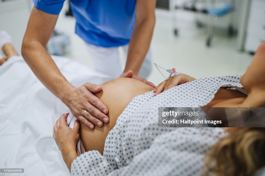 Obstetrician examining pregnant woman in hospital, touching woman’s belly, examine position of baby, breach position, size of baby and movements. Woman in labour laying on hospital bed, talking with ob-gyn doctor in maternity ward.