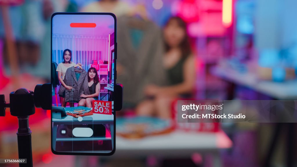 Closeup of young Asian women live streaming on phone show new collection fashion clothes to customer in shop at night. SME small business e-commerce.
