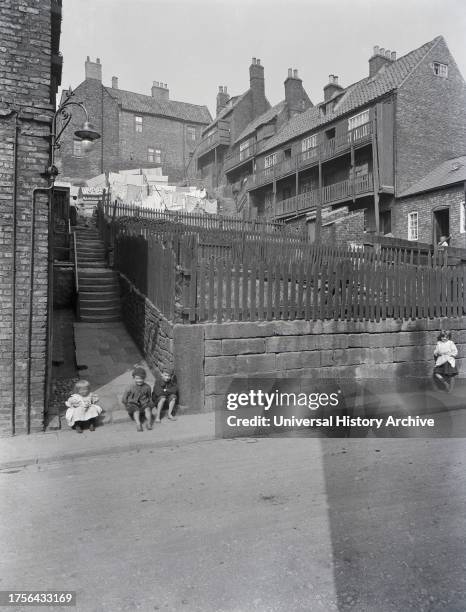 Glass negative 1900, Victorian era. A street in Whitby, Yorkshire with four children. Three of the children are bare foot. Above the street women are...