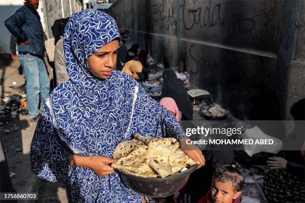 Girl walks carrying a pot filled with fresh traditional flatbread as women sit to bake more along an alley in Rafah in the southern Gaza Strip on...