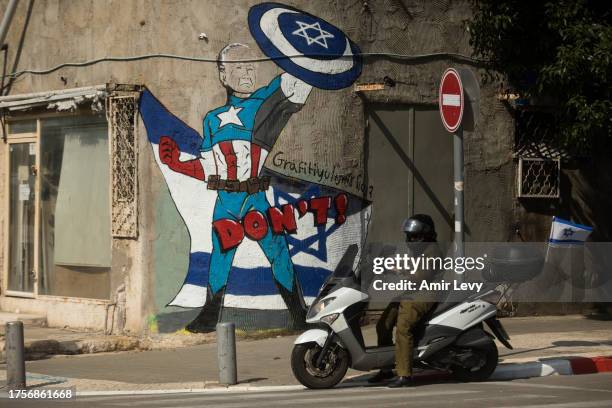 An Israeli soldier rides motorcycle near street art showing US President, Joe Biden, as Captain America super hero on a street in southern Tel Aviv...