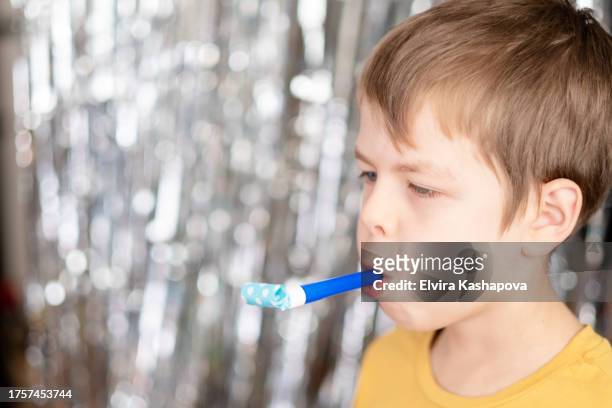 9-year-old boy in a yellow t-shirt blows a whistle at a birthday party against a background of silver sparkles. - blechblasinstrument stock-fotos und bilder