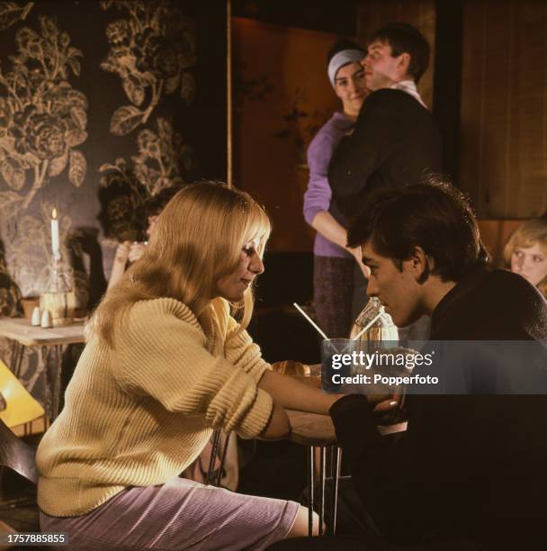 English art student Laura Harvey, seated on left wearing a yellow V-neck jumper, holding hands with a male friend as they share a glass of cola and a...