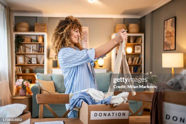 a caring young woman packs clothes for donation into a cardboard box - declutter stockfoto's en -beelden