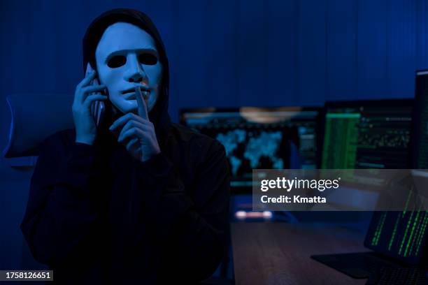 unidentified hacker using phone looking at the camera with finger on lips, asking for silence while working on computer crime. - anonymous mask photos et images de collection