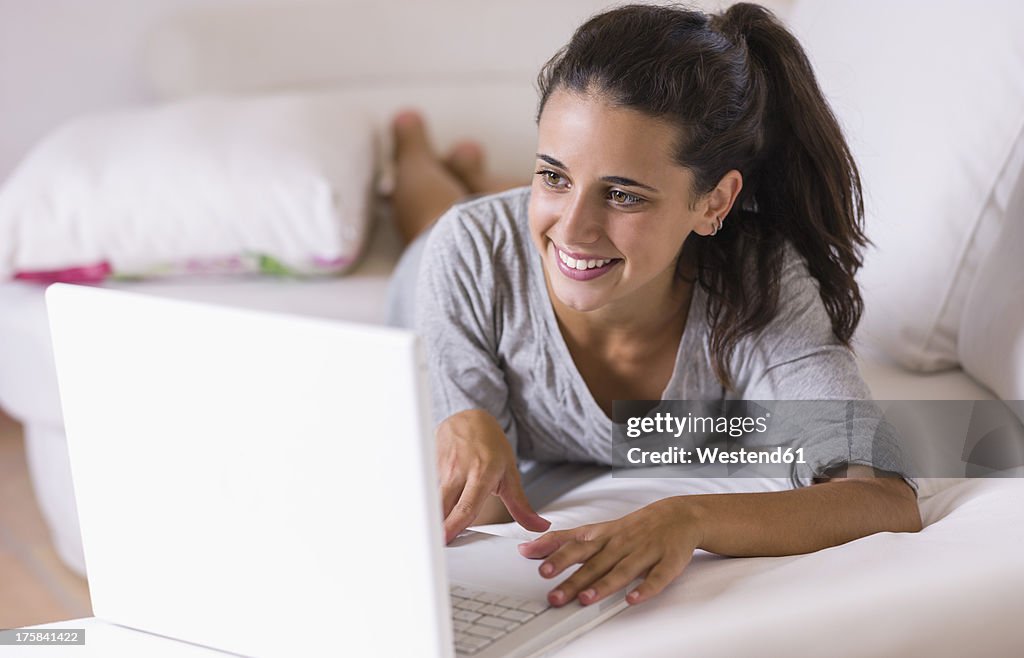 Teenage girl lying on white couch with laptop, smiling