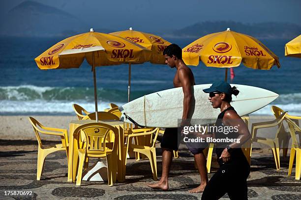 Surfer and a jogger at Ipanema beach pass by tables and umbrellas displaying the logo for Cia. De Bebidas das Americass Skol brand beer in Rio de...