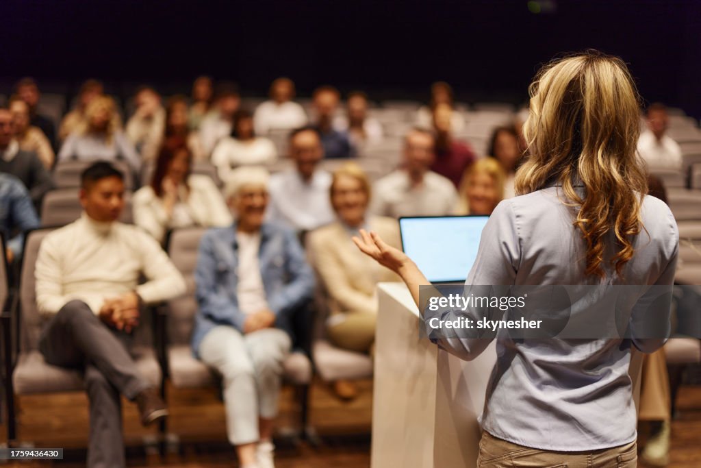 Back view of a female speaker giving a speech in front of people at convention center.