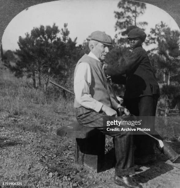 American businessman and philanthropist John D Rockefeller, his golf club resting across his lap as he sits on a small bench, his caddie standing to...