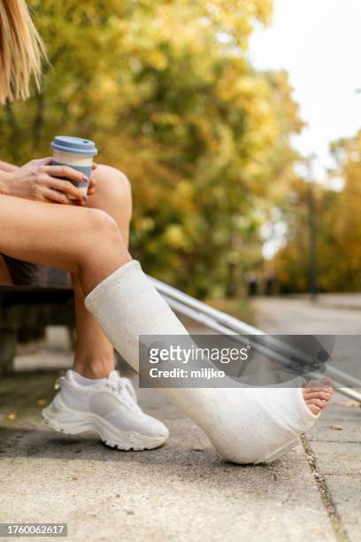 woman with a cast on her leg sitting on a bench in a park - woman in broken shoe heel stockfoto's en -beelden