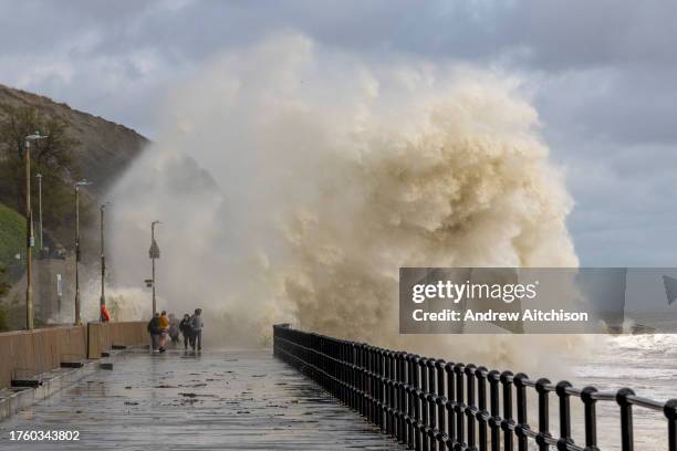 People walk along the sea defence promenade walkway while waves crash over them on Sunny Sands Beach, Folkestone, UK on the 2nd of November 2023 as...