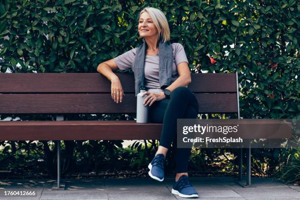 senior woman taking a break after her intense outdoors workout and sitting on the bench - slank stockfoto's en -beelden