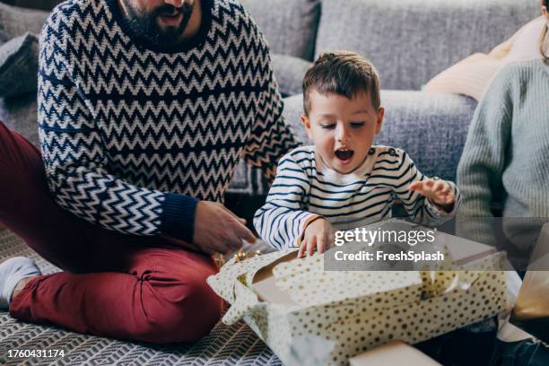 un lindo bebé abriendo su regalo de navidad mientras está sentado con sus padres irreconocibles en casa - desenvolver fotografías e imágenes de stock