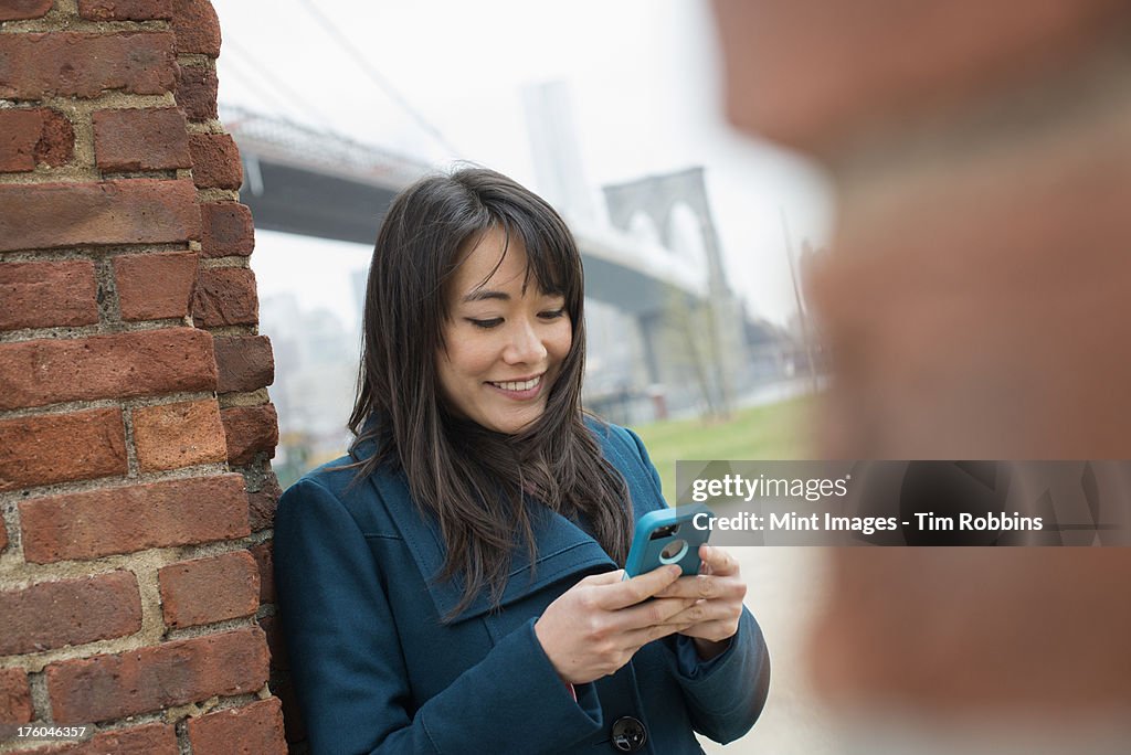 New York city. The Brooklyn Bridge crossing over the East River. A woman leaning against a brick wall, checking her phone.