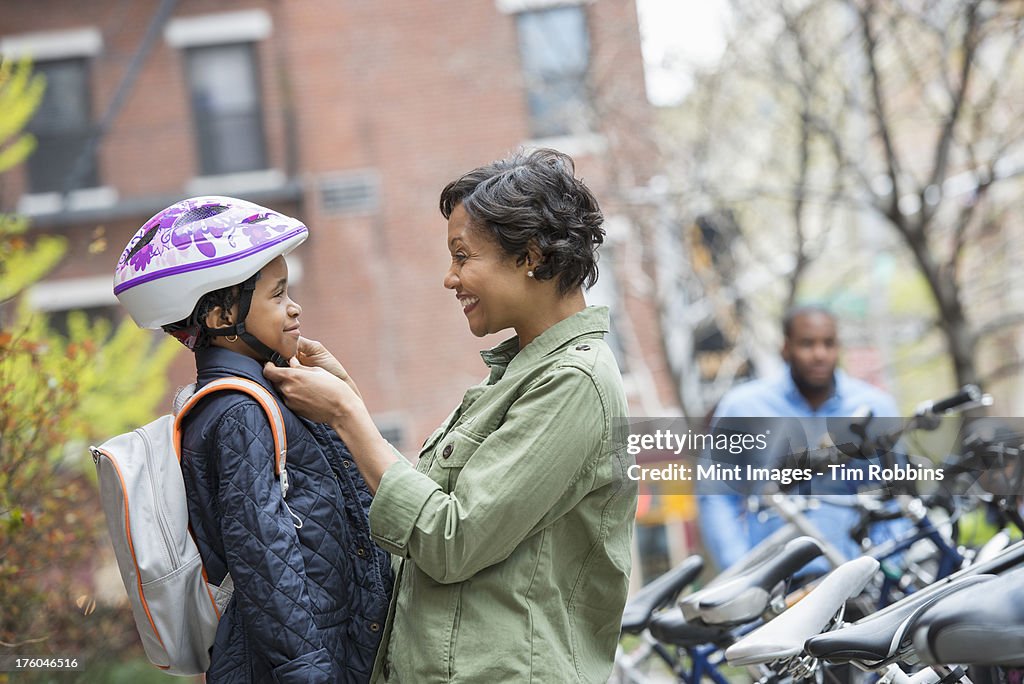 A New York city park in the spring. A boy in a cycle helmet, being fastened by his mother, beside a bicycle rack.