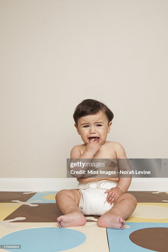 An 8 month old baby boy sitting on the floor in a diaper crying.