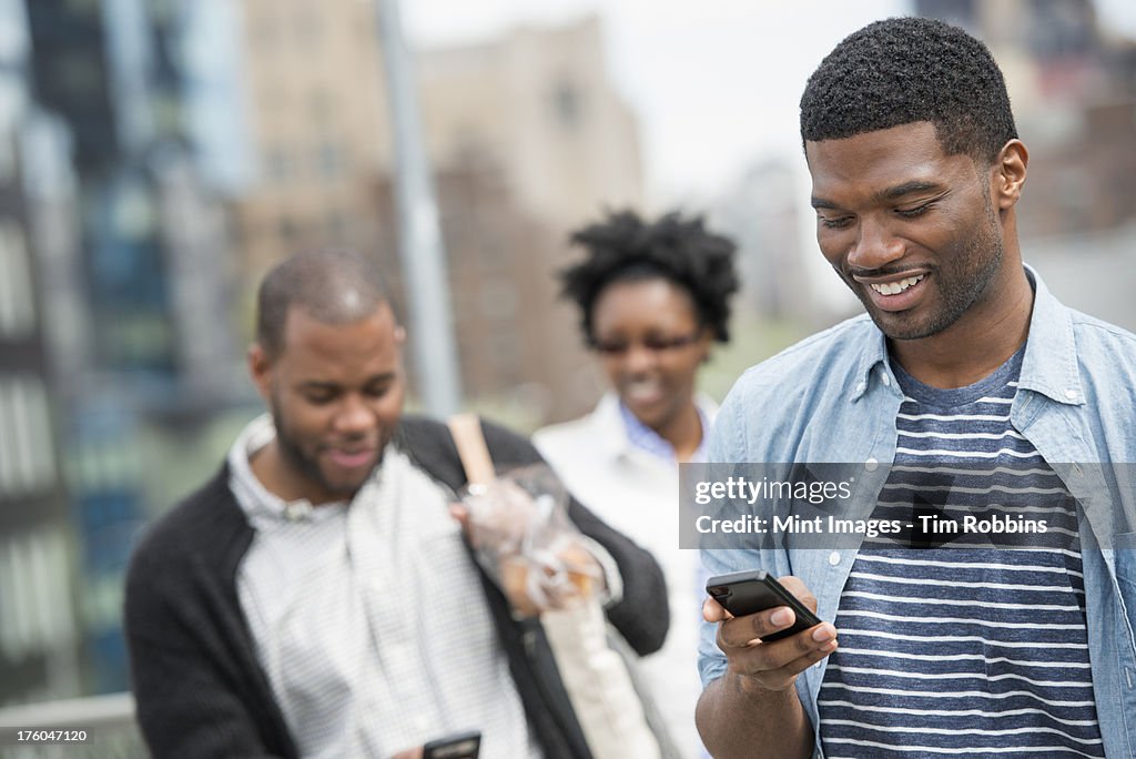 Outdoors in the city in spring. An urban lifestyle. A young man checking his phone and texting. A couple behind him.