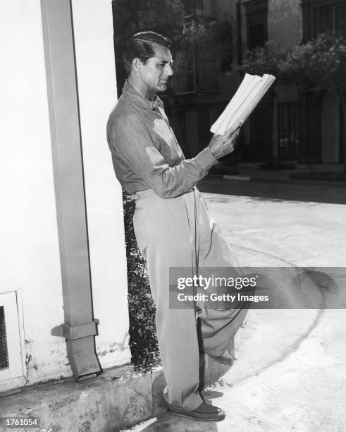 British born actor Cary Grant leans against a building while studying a script on a studio backlot, Los Angeles, California, 1955.