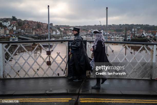 Couple wearing gothic style clothing walk across the swing bridge as they attend Whitby Goth Weekend on October 28, 2023 in Whitby, England. The...