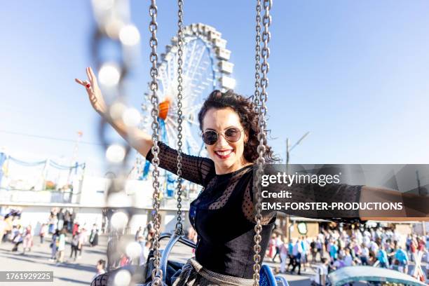 happy woman with long dark curly hair and sunglasses is wearing traditional bavarian dress while enjoying chain swing ride with giant wheel in background. - zweefmolen stockfoto's en -beelden