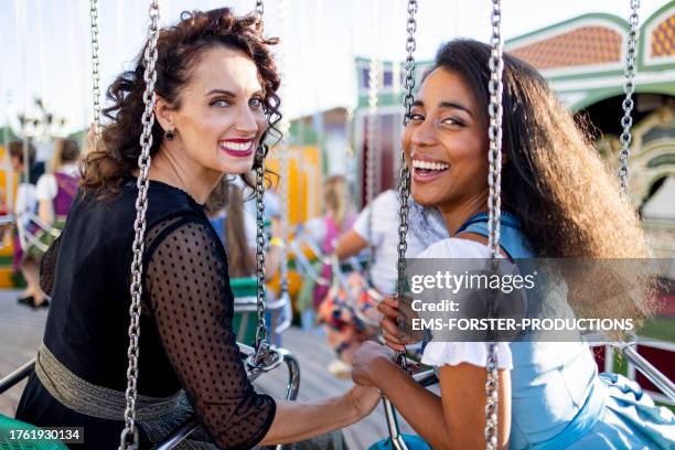 two female best friends of different age and race having great fun together in a chain swing at a beer festival in munich, they looking at camera and holding hands. - dirndl stock pictures, royalty-free photos & images