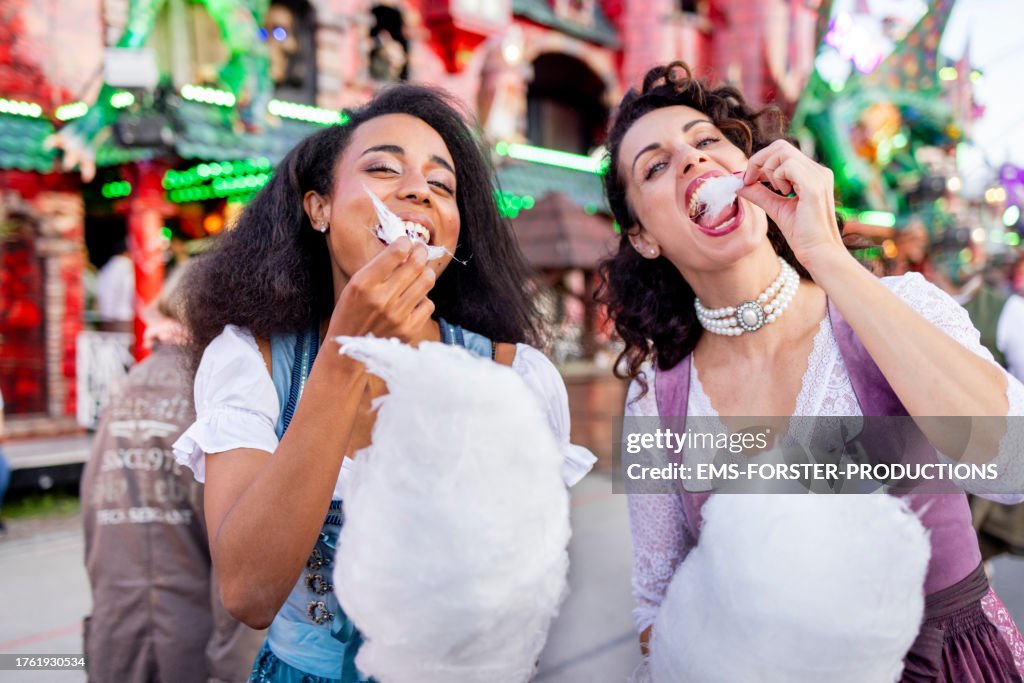 Female best friends in traditional Bavarian clothing eating candy floss while walking at a beer festival in Munich and looking at camera.
