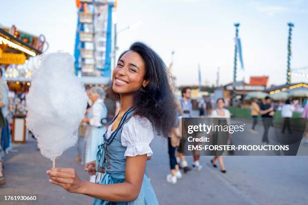 woman of mixed race wearing traditional bavarian dirndl dress holds stick with cotton candy while walking at a beer festival in munich and looking at camera. - bierfest stock-fotos und bilder