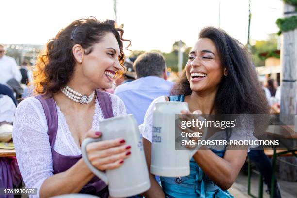 two female best friends of different age and race having great fun together at a beer festia; in munich while drinking beer outdoors in the garden. - biergarten münchen stock pictures, royalty-free photos & images