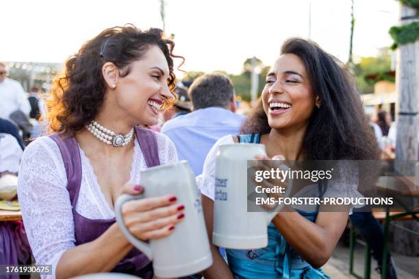 two female best friends of different age and race having great fun together at a beer festia; in munich while drinking beer outdoors in the garden. - saia tipo camponesa com ou sem corpete - fotografias e filmes do acervo