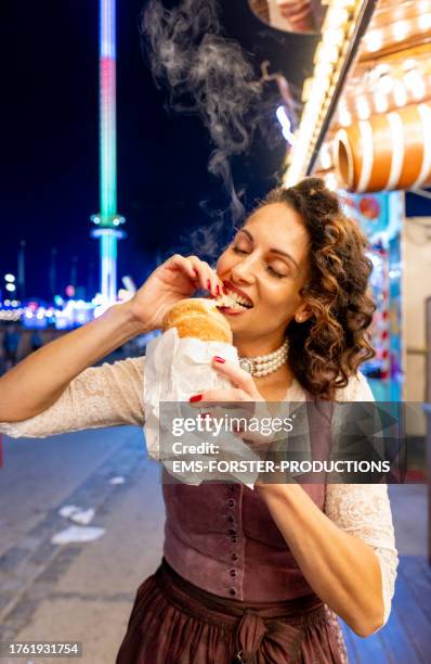 mature beauty woman wearing traditional bavarian dirndl dress is eating hot bakery snack outdoors at beer festival in munich while night. - saia tipo camponesa com ou sem corpete - fotografias e filmes do acervo