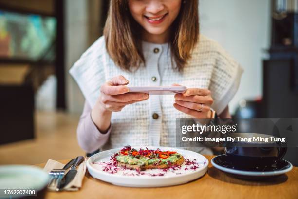 young cheerful asian woman taking photo of her delicious breakfast with smartphone in a restaurant - foodie stockfoto's en -beelden