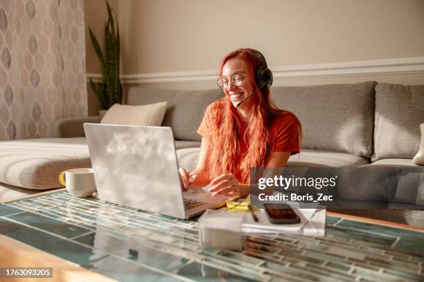 a beautiful young woman with red hair and glasses is working from home, multitasking. she's on an online team meeting with headphones. - associations icon stock pictures, royalty-free photos & images