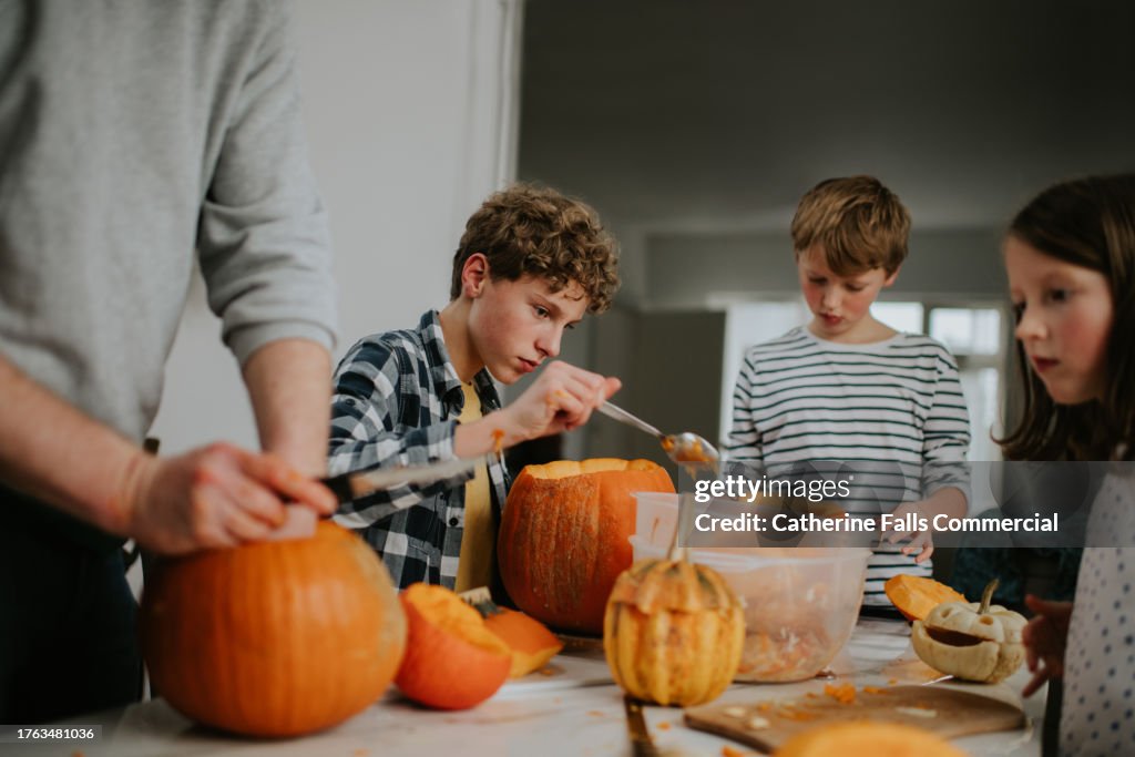 Three children and an adult sit around a table in a domestic environment and make Jack O' Lanterns out of large pumpkins