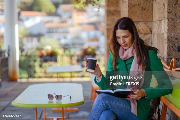 woman reading a book and drinking coffee sitting at a table in a cafe - lerexis stockfoto's en -beelden